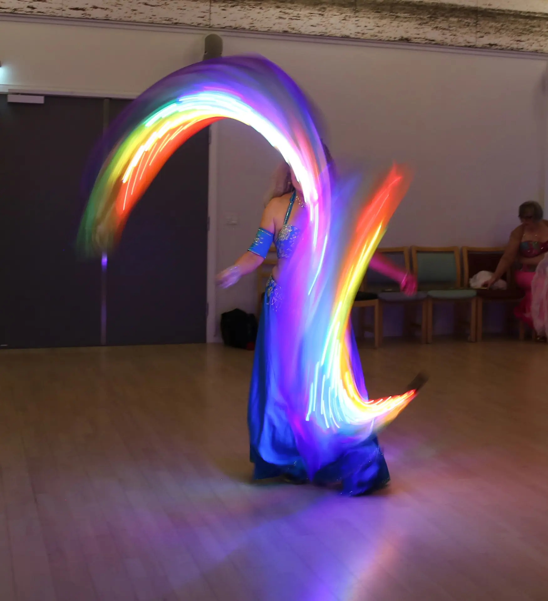 Aziza svinger to silke‑poi‑slør i bevægelse, iført et regnbuefarvet kostume.