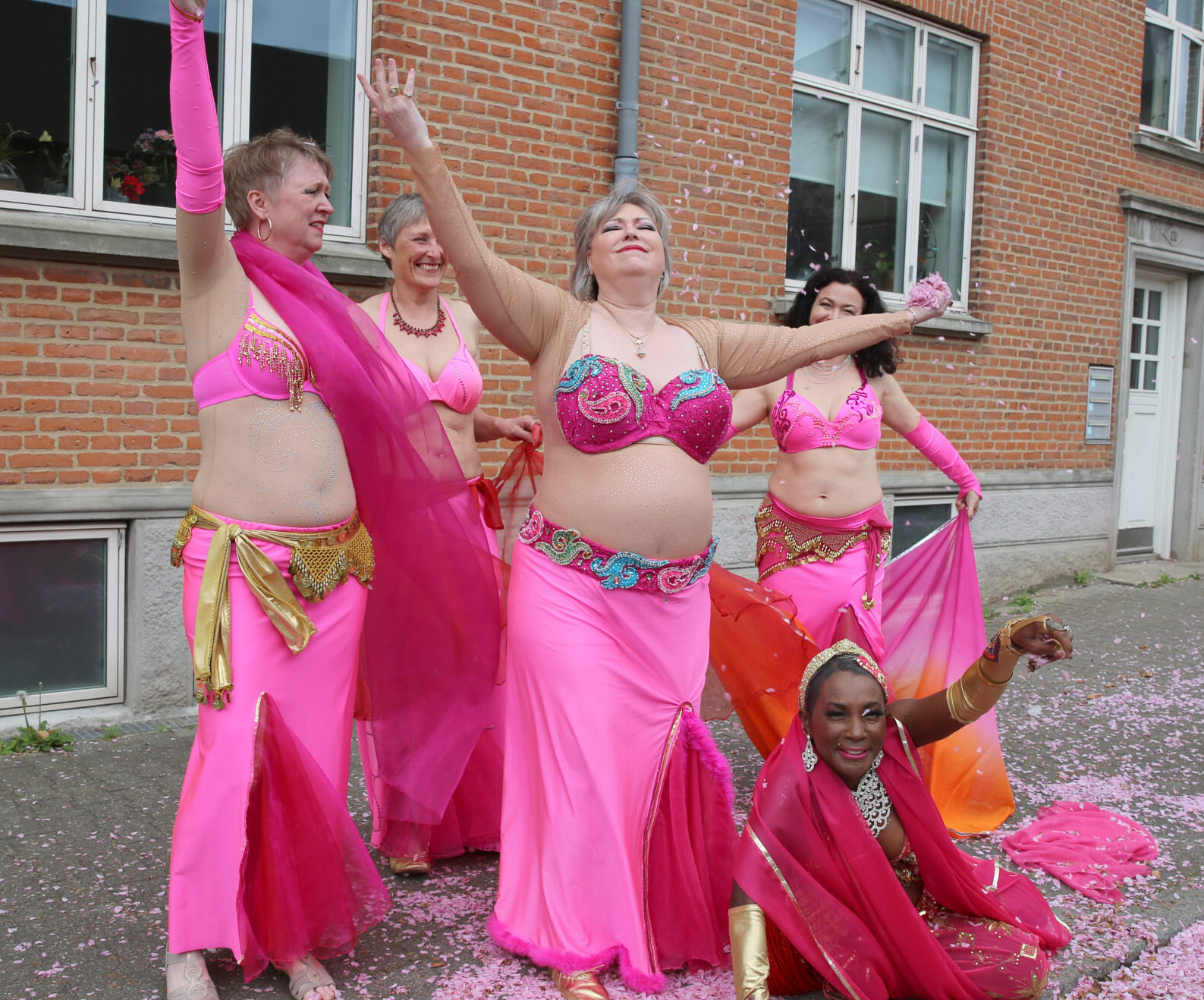 Dansere fra Ensemble Amira i pink kostumer kaster lyserøde kirsebærblomster i luften under fotosessionen 2025. Nogle står, andre sidder eller ligger på gaden, omgivet af blomster.