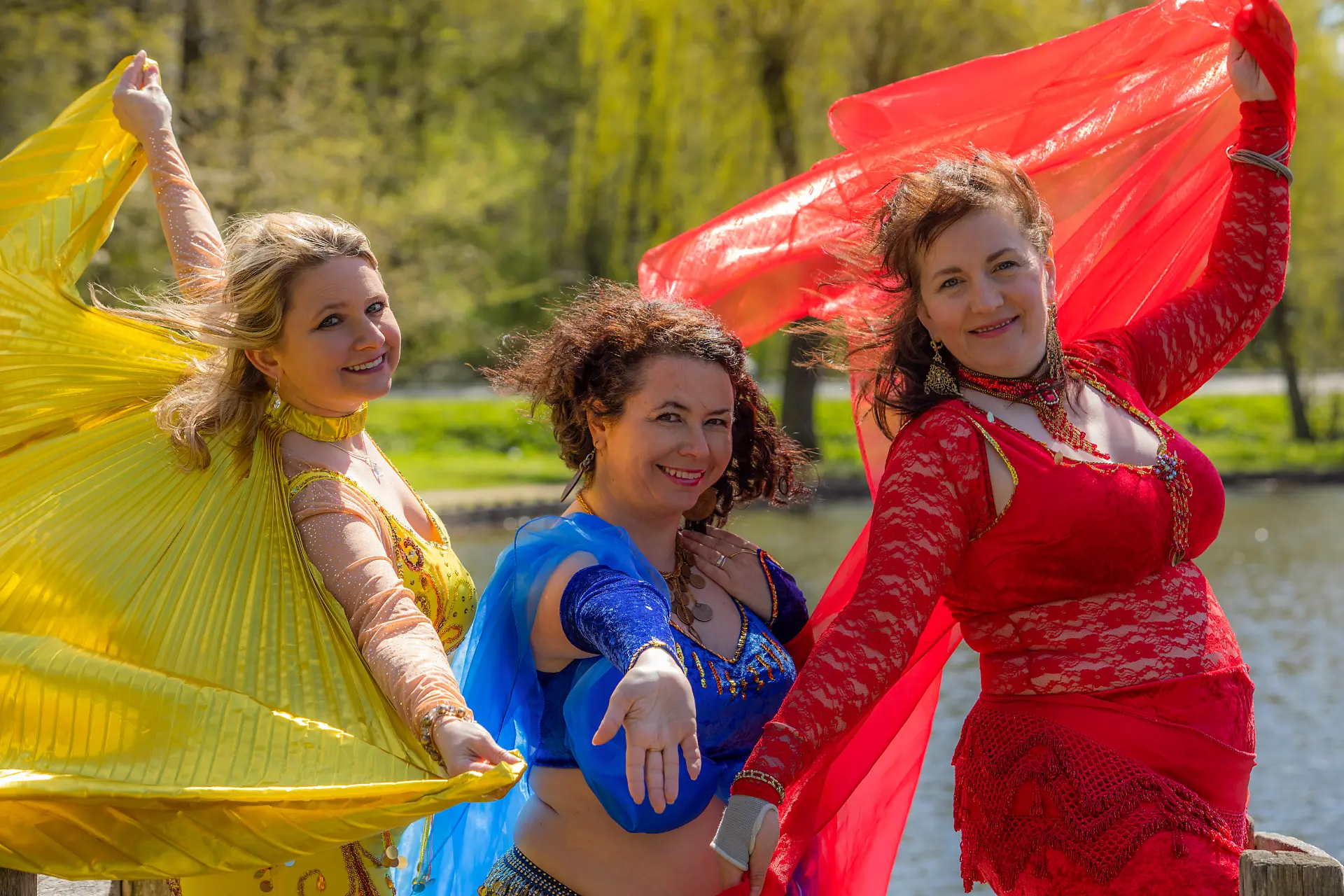 Tre kvinder i farverige mavedans‑kostumer i gul, blå og rød står i en park og rækker hånden ud mod beskueren i en inviterende bevægelse. Foto: Thomas Eriksen.